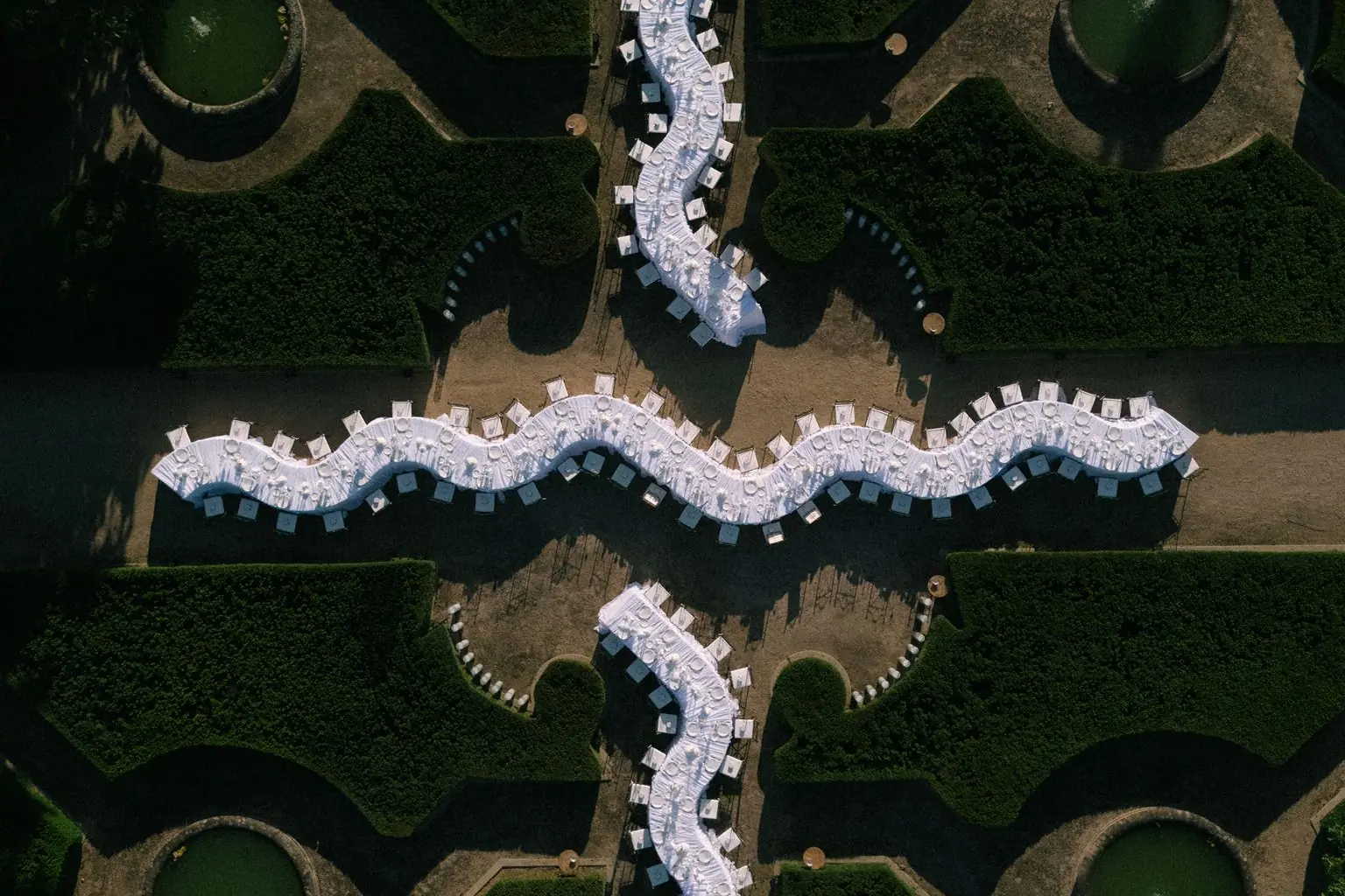 Vista desde el aire de un jardín de boj con tres mesas largas y onduladas, vestidas de blanco, que forman una cruz